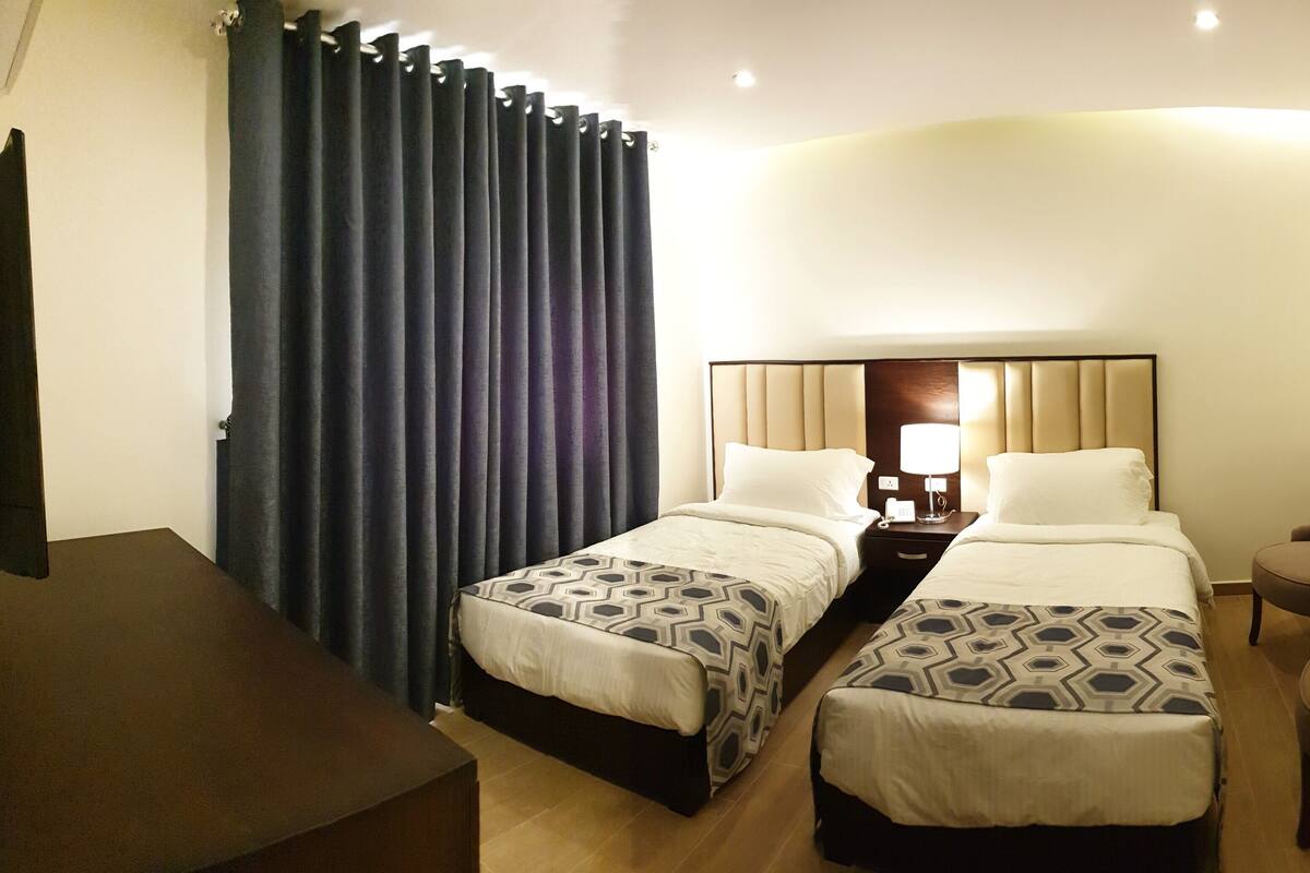 A hotel room with two beds, a desk, a chair, and a large window with curtains.