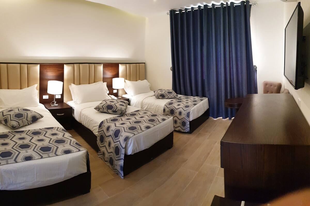 A hotel room with three beds, each with patterned bedding, a wooden headboard, bedside lamps, and a large window with dark curtains.