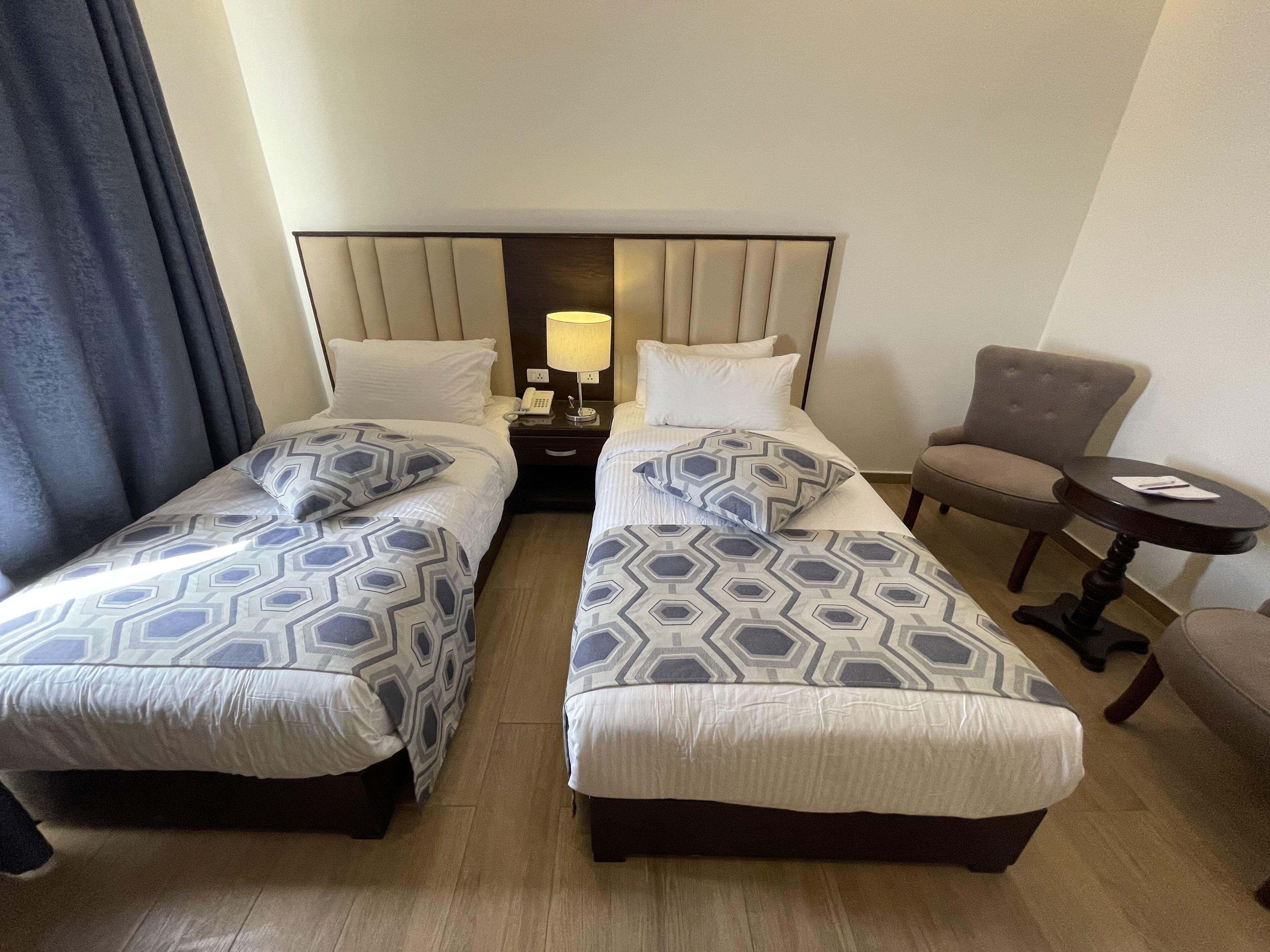 A hotel room with two beds, a nightstand with a lamp, a chair, and a small table.