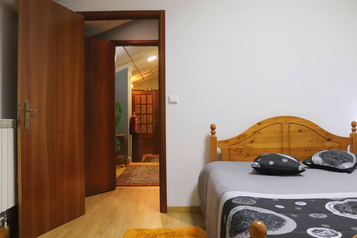 Quarto com cama de madeira, mesa de cabeceira, tapete pequeno e porta que dá para outro cômodo.