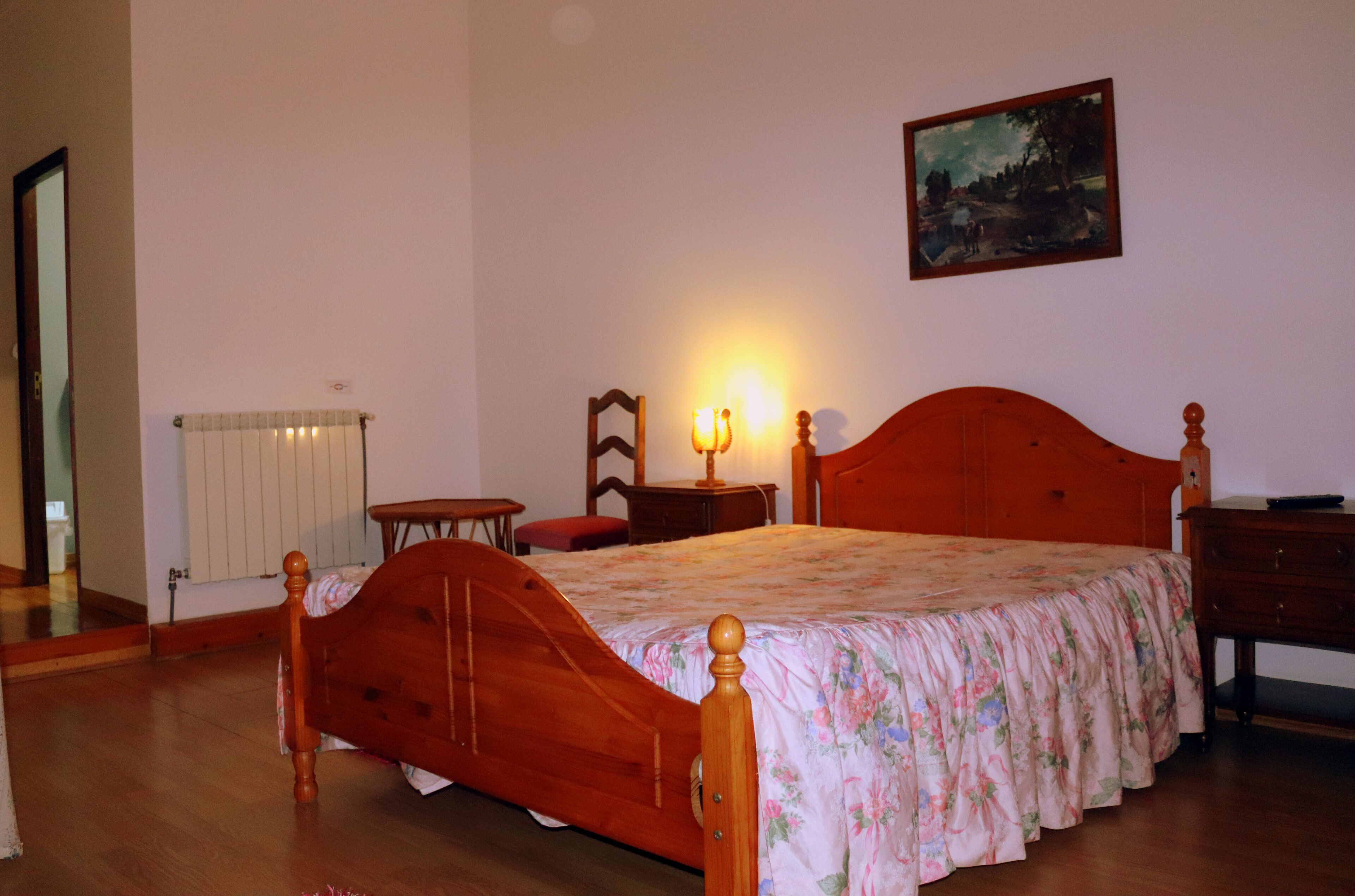 Une chambre avec un lit en bois, une table de chevet, une commode, une chaise, un radiateur et un tableau accroché au mur.