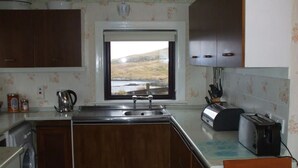 A compact kitchen with a window, wooden cabinets, a sink, a kettle, a toaster, and a washing machine. - Private  Croft House in the Western Isles 
STL Licence Number ES00069F (Scotland)