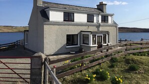 A two-story house with a grey exterior, a front porch, and a wooden fence. - Private  Croft House in the Western Isles 
STL Licence Number ES00069F (Scotland)