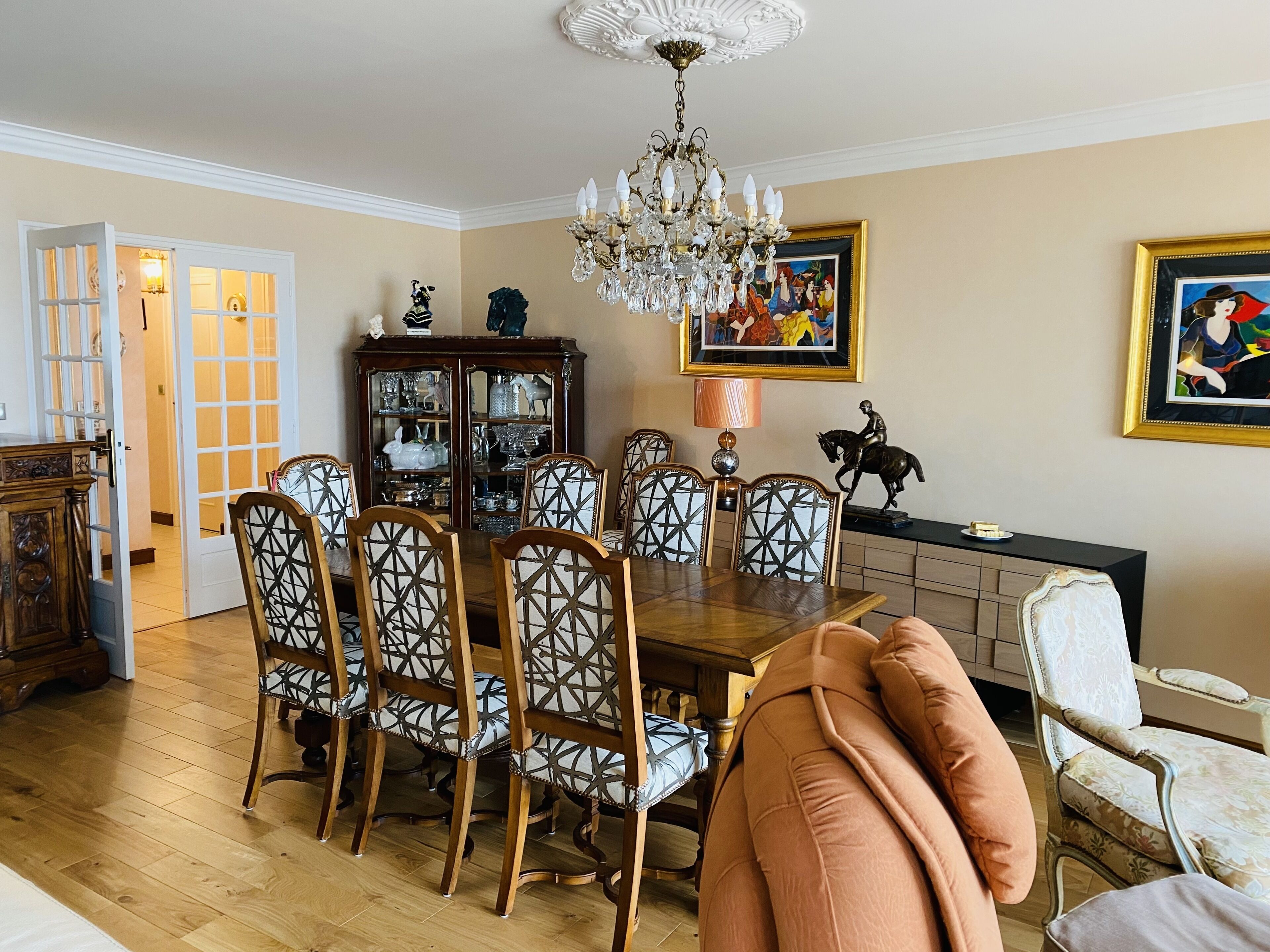 A dining room with a wooden table, six chairs, a chandelier, a cabinet with glass doors, and two framed paintings.
