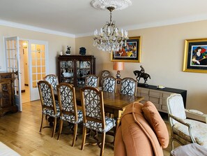A dining room with a wooden table, six chairs, a chandelier, a cabinet with glass doors, and two framed paintings.