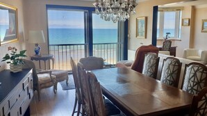 A spacious living room with a large wooden dining table, comfortable seating, and a view of the sea through large windows.