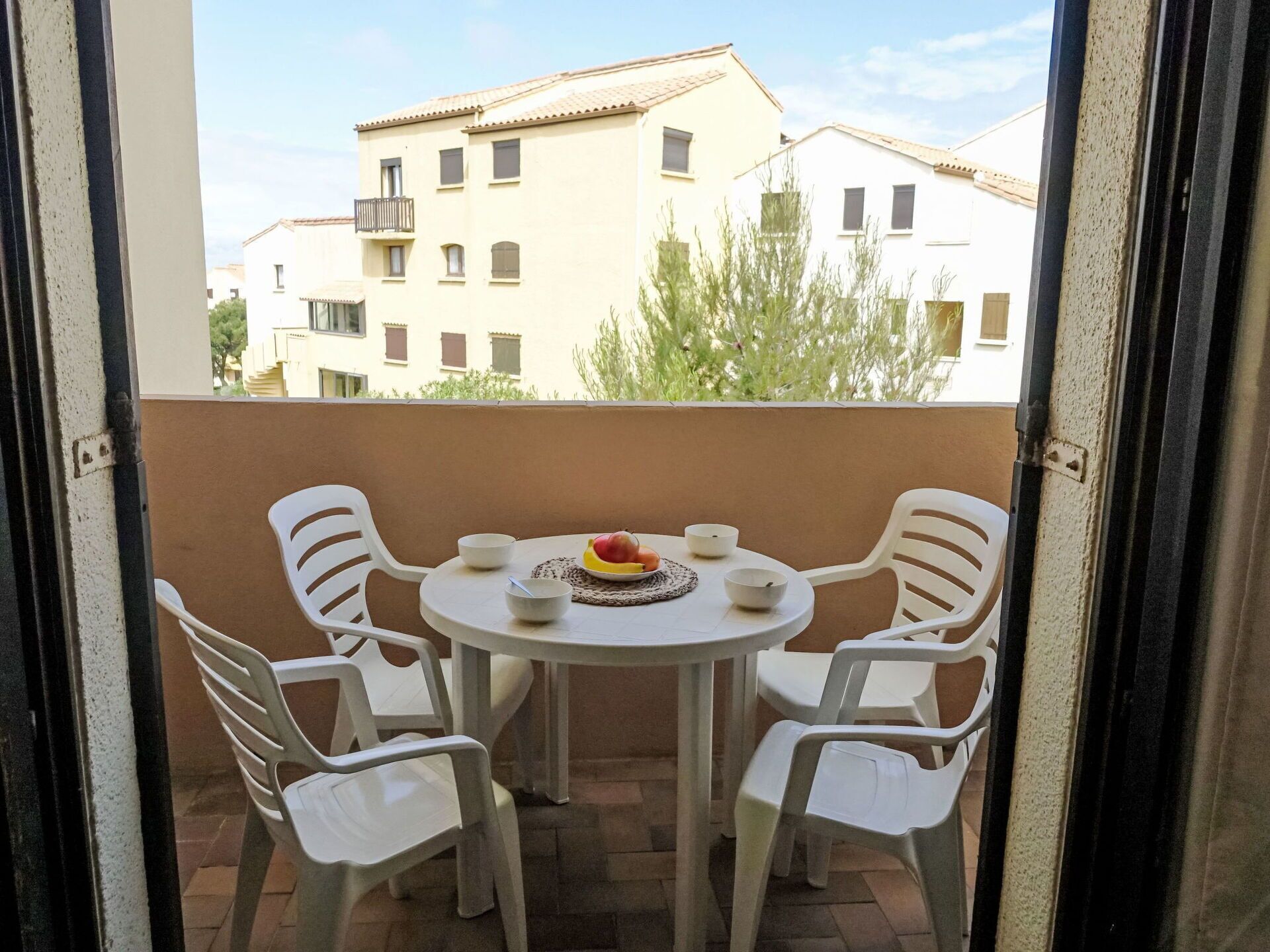 Un balcon avec une table ronde et quatre chaises en plastique blanc. Sur la table, on trouve une assiette avec des fruits et un bol.