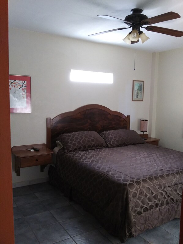 Un dormitorio con cama, mesita de noche, ventilador de techo y una lámpara en la pared.