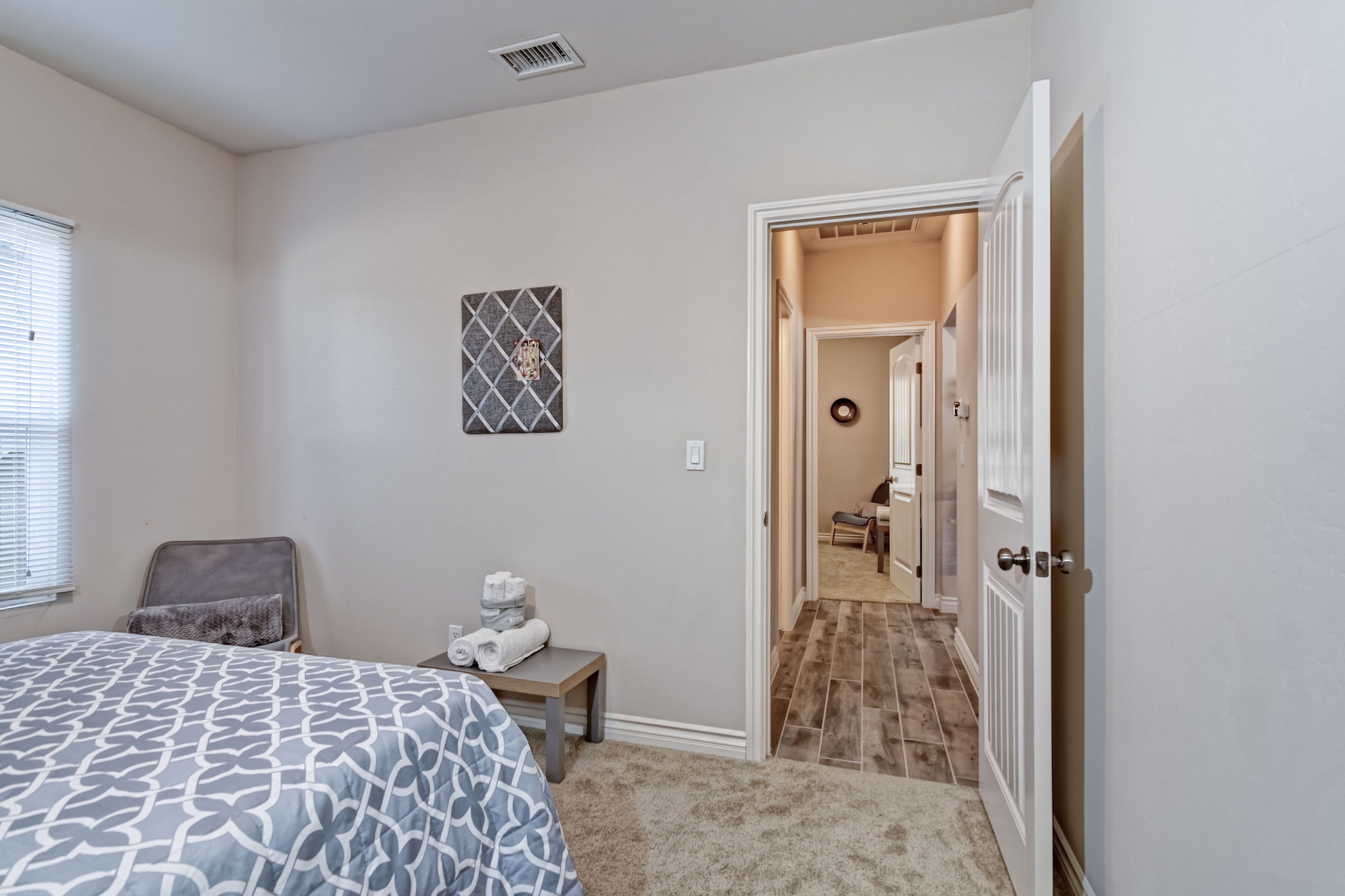 A bedroom with a bed, a small table with towels, a chair, a picture on the wall, and a door leading to a hallway.