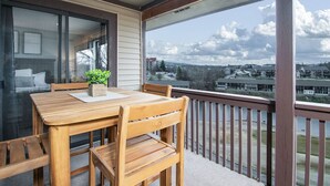 Terraza de madera con una mesa, sillas, una planta en maceta y vistas a edificios y árboles.
