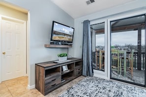 A living room with a flat-screen TV on a wooden stand, a sliding glass door leading to a balcony, and a patterned rug on the floor.