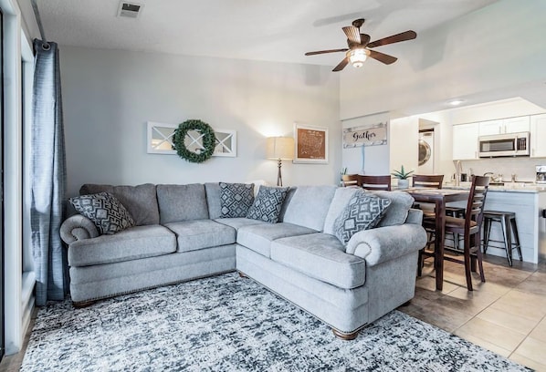A modern living room with a grey sectional sofa, a patterned rug, a ceiling fan, and a kitchen area in the background.