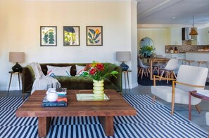 Een moderne woonkamer met een groene bank, een houten salontafel en een gestreept vloerkleed. Drie ingelijste kunstwerken hangen aan de muur en er is een eethoek met stoelen en een tafel.