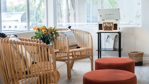 A room with wicker chairs, a red ottoman, a radio, and a window with a view of palm trees and a grill outside.