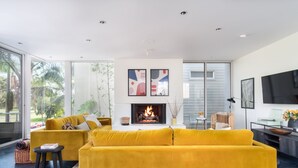 Ein modernes Wohnzimmer mit einem gelben Sofa, einem Kamin und großen Fenstern mit Blick ins Freie.