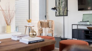 Ein modernes Wohnzimmer mit Couchtisch aus Holz, Büchern, einem Korbstuhl, einer Stehlampe und einem Fernseher auf einem schwarzen Standfuß.