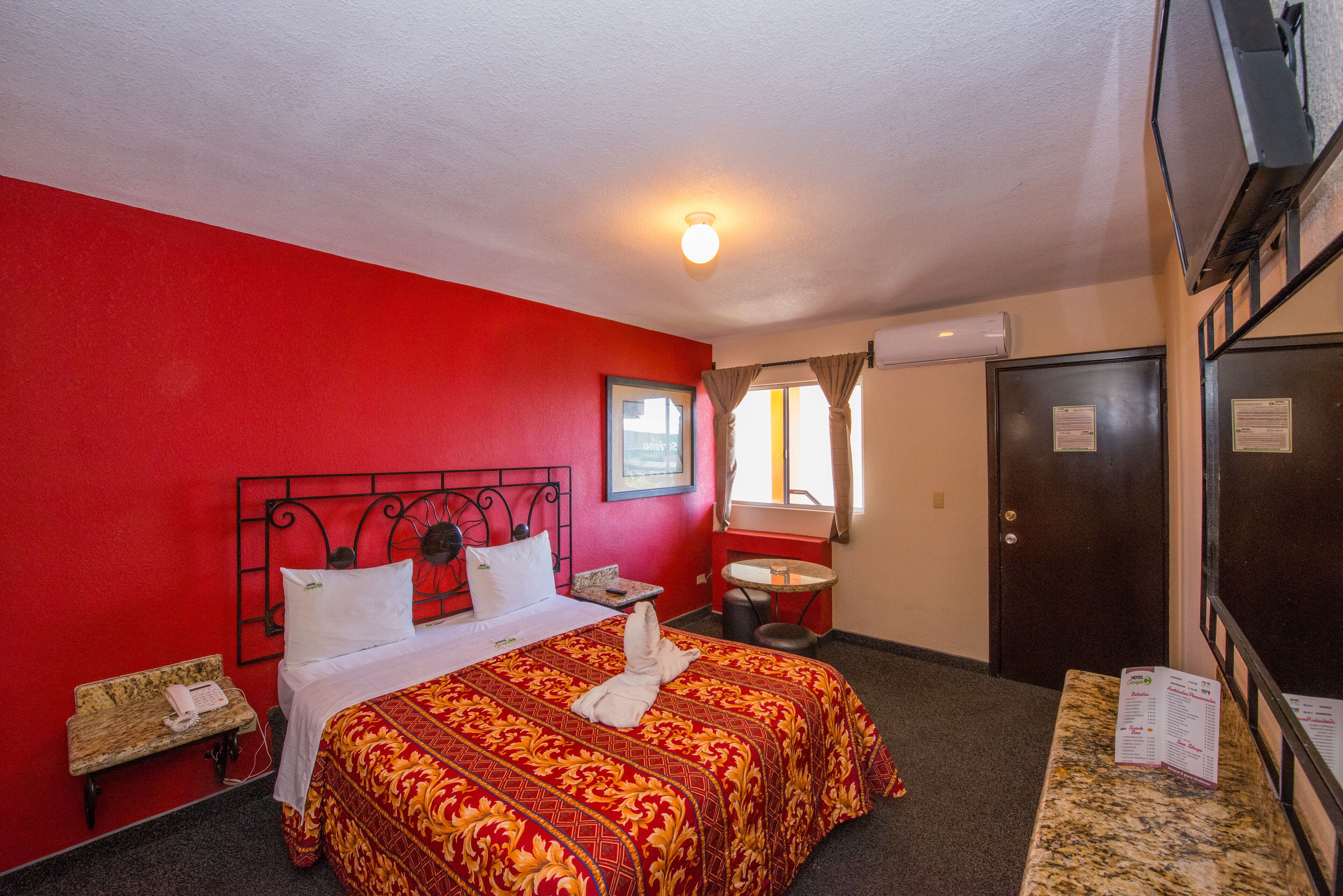 A hotel room with a red wall, a bed with a patterned bedspread, a bedside table with a lamp, a small round table, and a mirror.