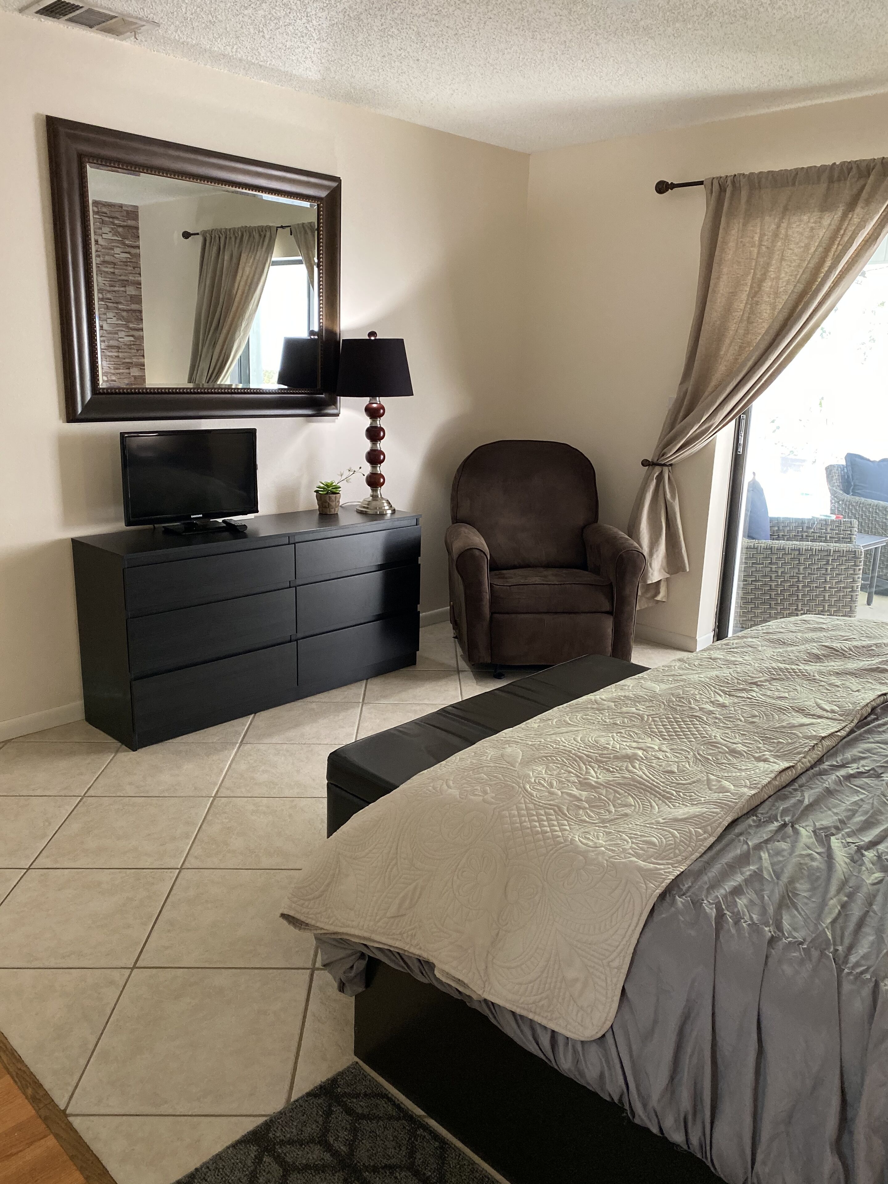Een slaapkamer met een bed, een dressoir, een stoel, een televisie en een raam met gordijnen.