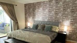 Une chambre moderne avec un grand lit, un ventilateur de plafond, un mur décoré d’un motif imitant la pierre et une vue sur l’extérieur.