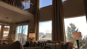 A spacious living room with a high ceiling, large windows, a balcony, and a dining area.