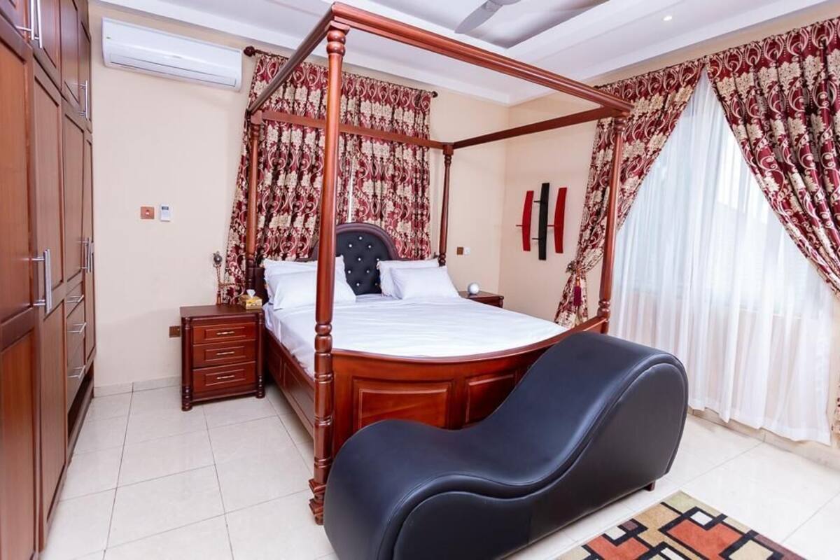 Uma cama com dossel e estrutura de madeira, uma mesa de cabeceira, uma poltrona estofada preta e um tapete estampado.