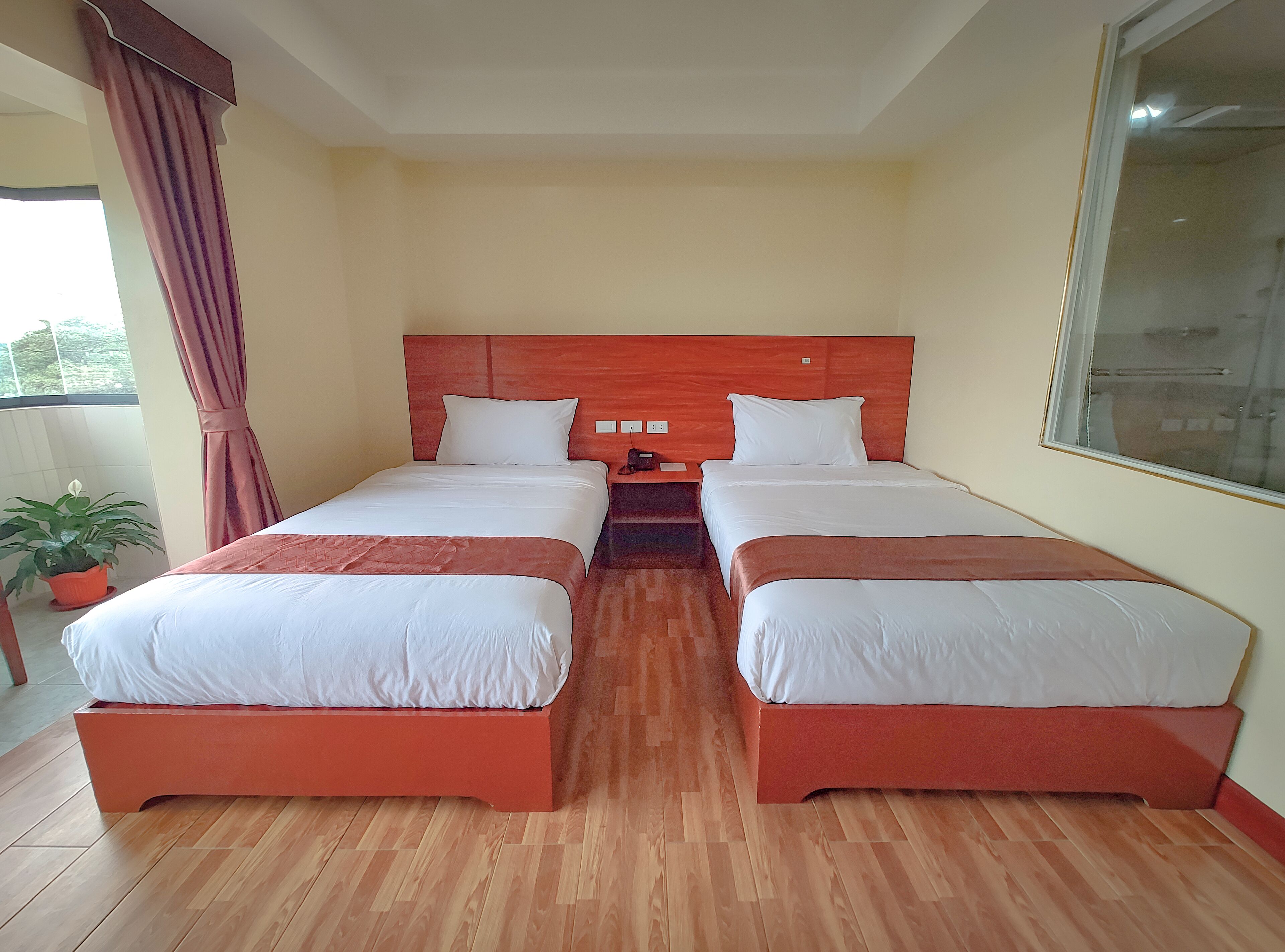 Camera d'albergo con due letti, una testiera in legno, un comodino, una pianta in vaso e una finestra con tende.