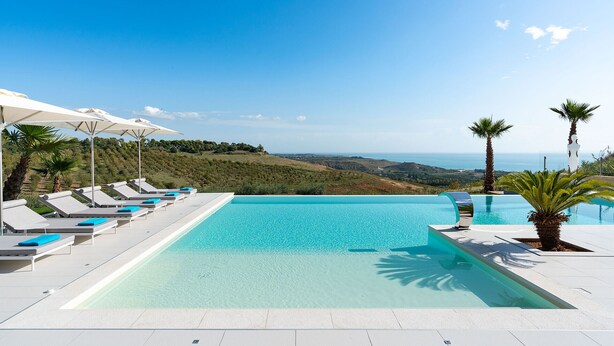 A pool with clear blue water, surrounded by white lounge chairs and umbrellas, with a view of the sea and hills in the background. - Luxury villa with pool situated near Ribera, Agrigento, Sicily – 6 pax (Ribera)