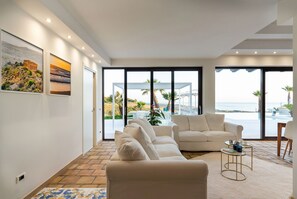 A modern living room with a beige sofa, a coffee table, and a view of the beach through large windows.