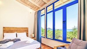 A bedroom with a wooden headboard, a bed with white linens, a grey blanket, and a view of greenery and water through large blue-framed windows. - 3 Bedroom Villa, Agios Sostis, Zakynthos (Lithakia)