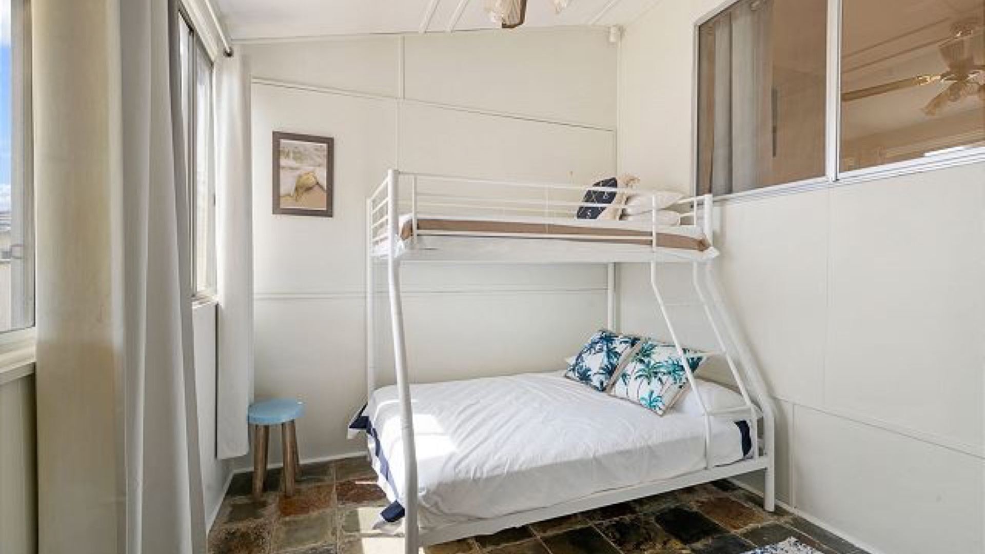 A bunk bed with a small stool, a framed picture on the wall, and a window with curtains.