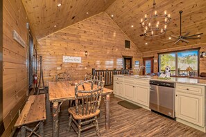 Ein Holzblockhaus-Interieur mit Esstisch, Stühlen, Küchenzeile und Blick ins Freie.