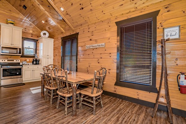 A rustic cabin interior with a wooden dining table, chairs, a kitchen with appliances, and a large window with blinds. - ⛰️ Awesome Star Splash 🌄 Indoor Pool, Hot Tub, Home Theater & Game Room (Sevierville)
