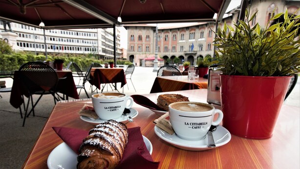 Ein Café im Freien mit Tischen, Stühlen und Blick auf Gebäude im Hintergrund.