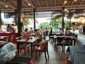 An outdoor dining area with multiple tables and chairs, a counter with a person serving, and a view of greenery outside. - PP Mountain Beach Resort (Ko Phi Phi)
