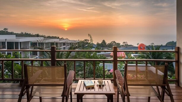 A wooden deck with two chairs and a table, overlooking a scenic sunset view with buildings and greenery in the background.