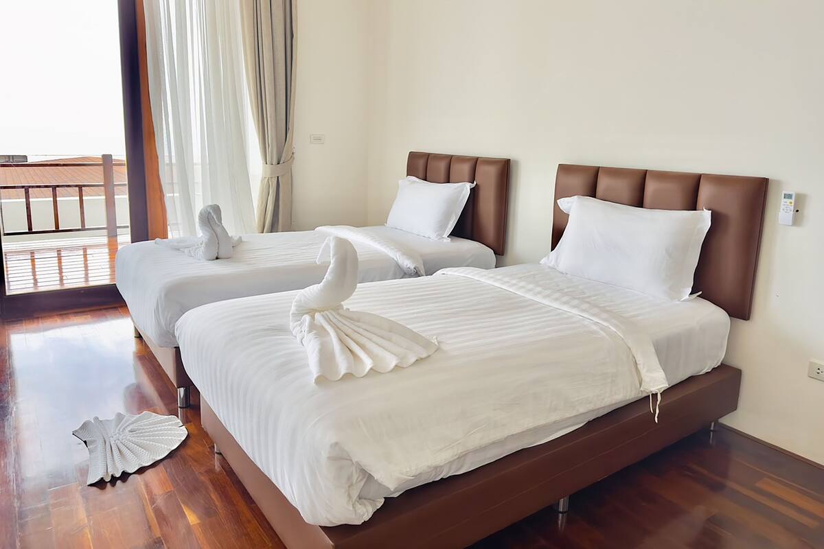Habitación de hotel con dos camas, cada una con cabecero marrón y ropa de cama blanca. El suelo es de madera y se ve un balcón por la ventana.