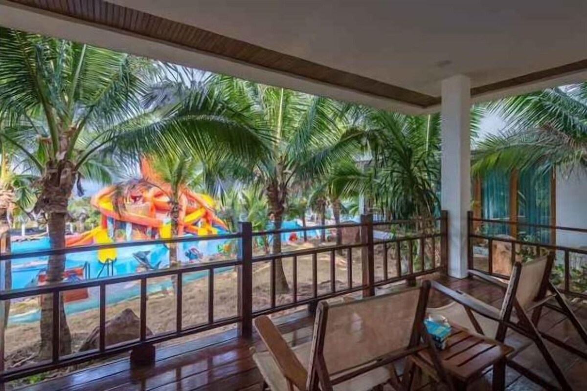 A covered outdoor area with wooden flooring, seating, and a view of a water park with slides and palm trees.