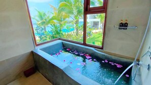A bathroom with a large window, a bathtub with water and flowers, and a shower area.
