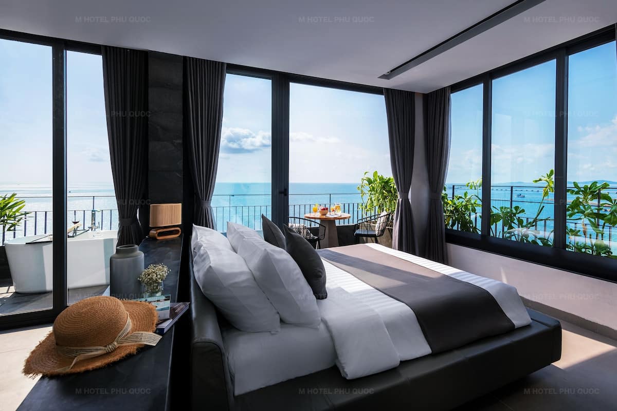 A modern bedroom with a large bed, a balcony with a view of the sea, and a dining area with a table and chairs.