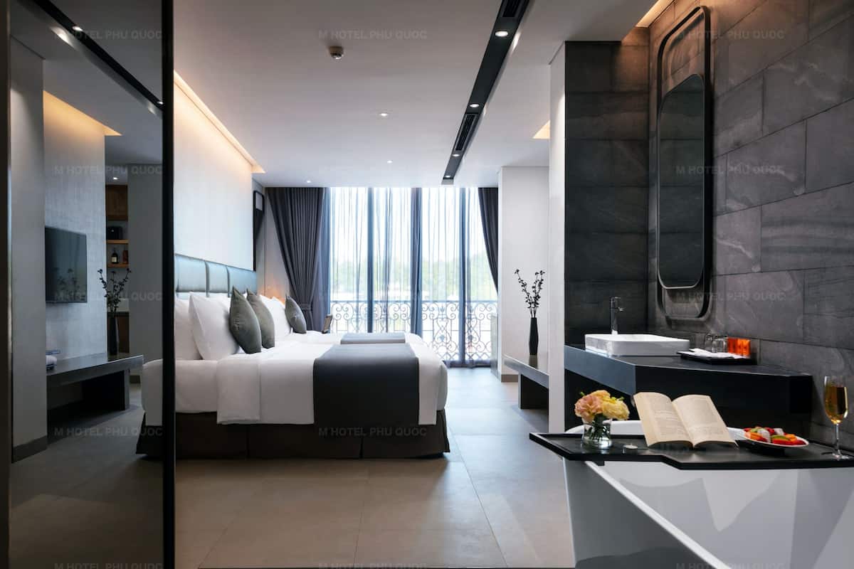 A modern hotel room with a large bed, a bedside table with a book and a glass of wine, a bathroom with a mirror, and a view of the outdoors through large windows.
