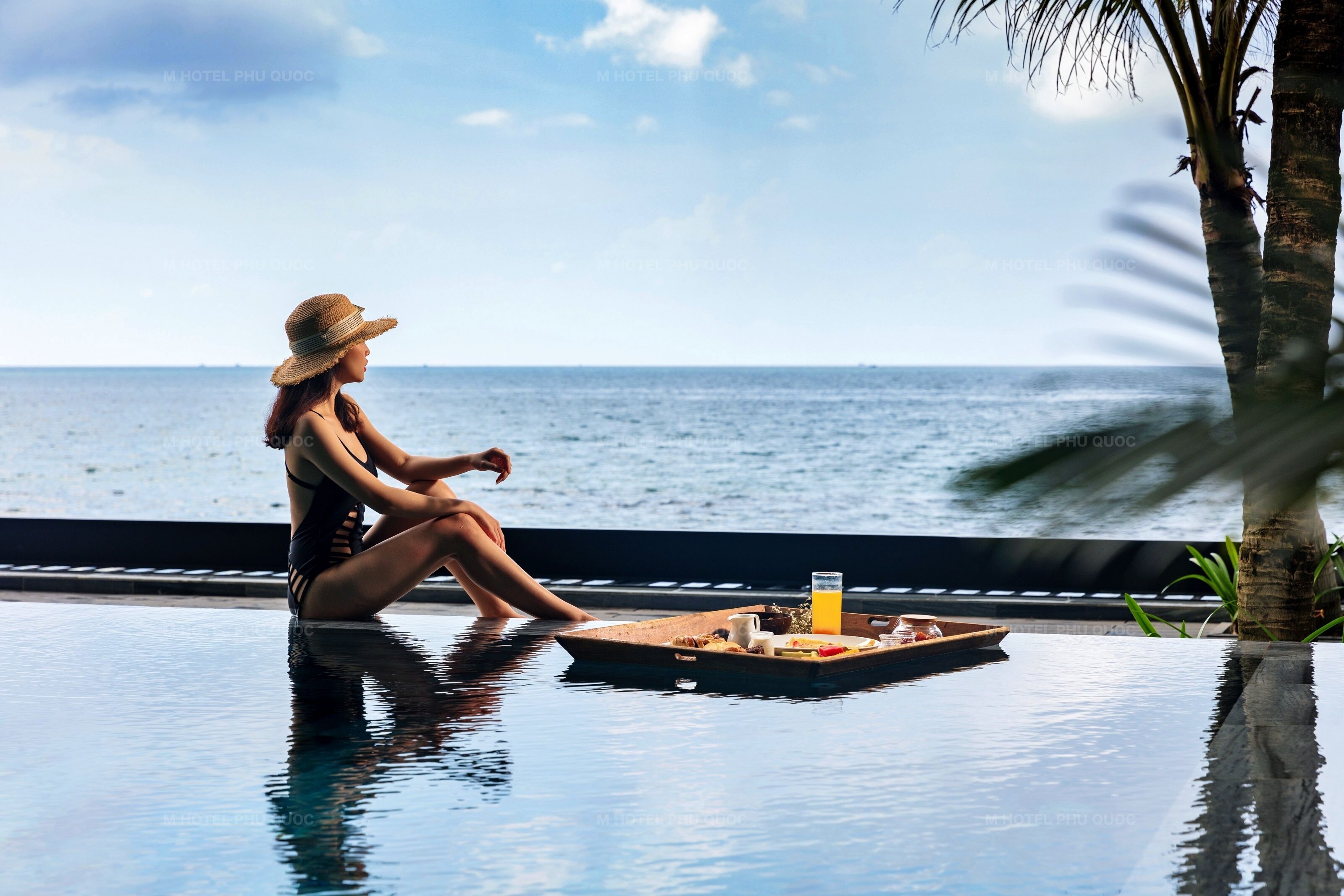 A woman sitting by a poolside table with a tray of food and drink, overlooking the ocean.