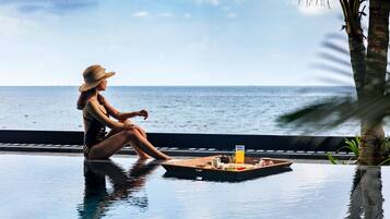A woman sitting by a poolside table with a tray of food and drink, overlooking the ocean.