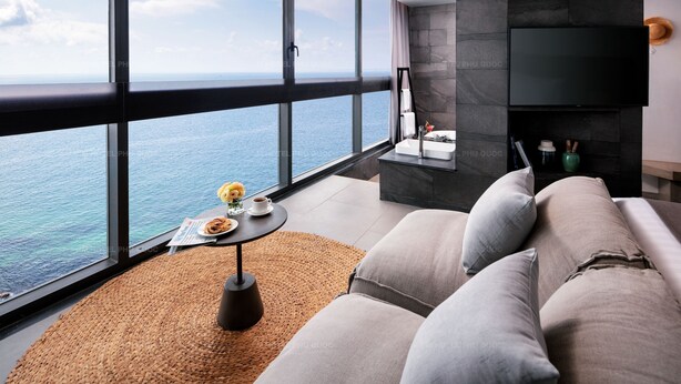 Ein modernes Wohnzimmer mit großem Fenster mit Meerblick, einer Couch, einem kleinen runden Tisch mit Teller und Tasse sowie einem an der Wand befestigten Flachbildfernseher.