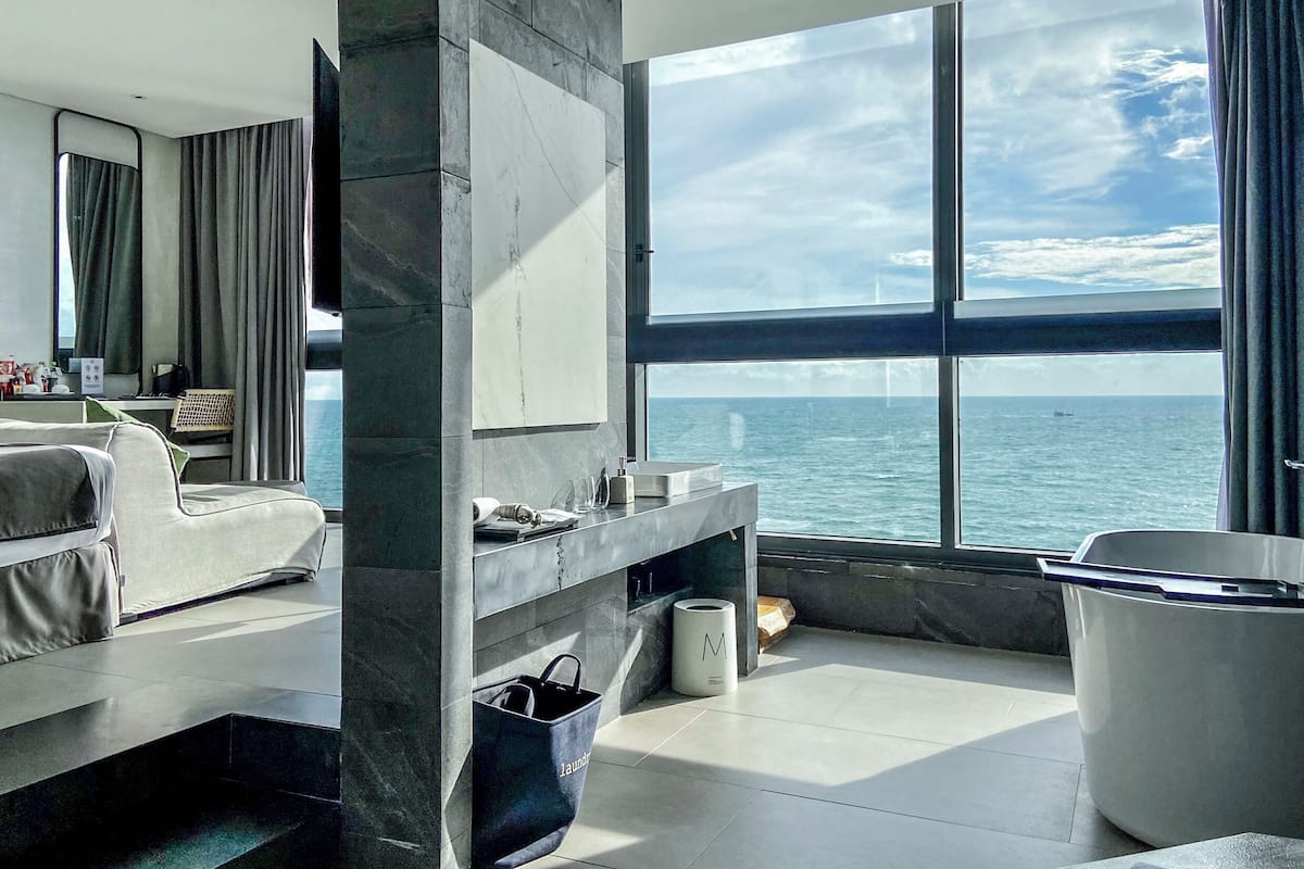 A modern hotel room with a large window offering a sea view, a sofa, a vanity with toiletries, and a trash bin.