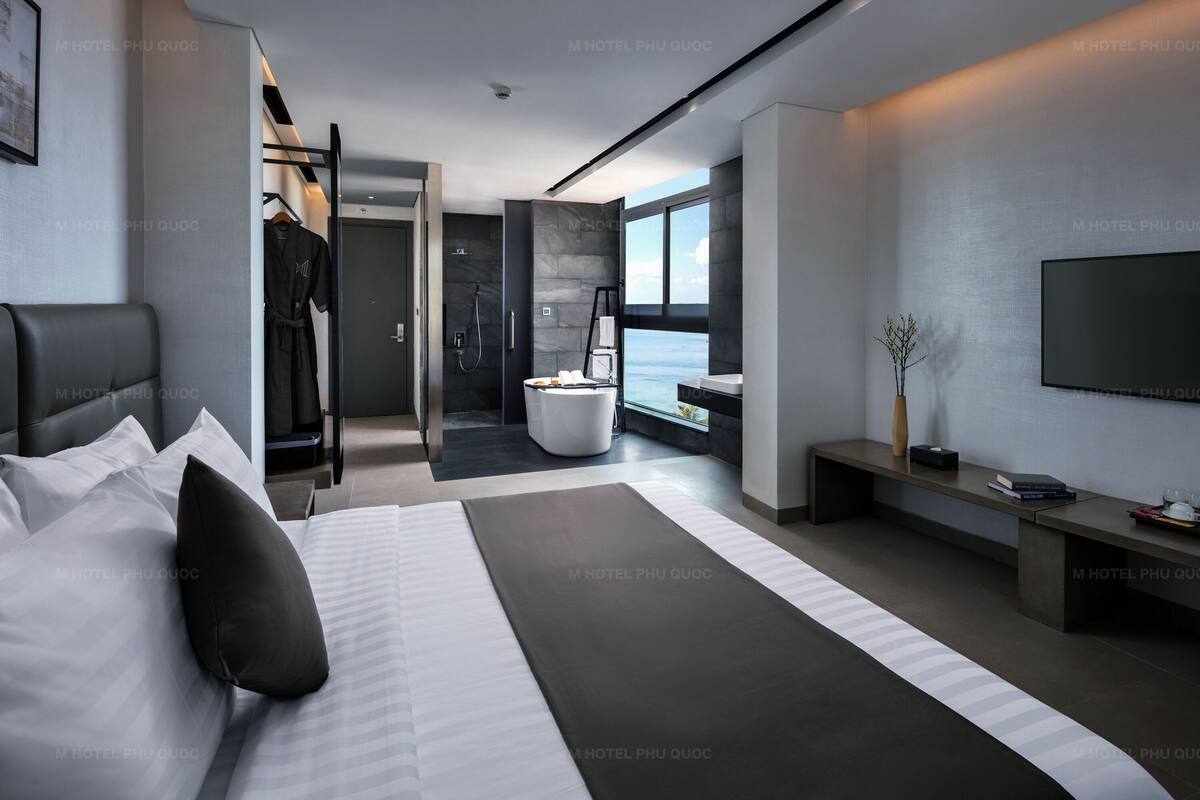 A modern hotel room with a large bed, a walk-in closet, a bathroom with a shower and bathtub, a TV, and a view of the sea.