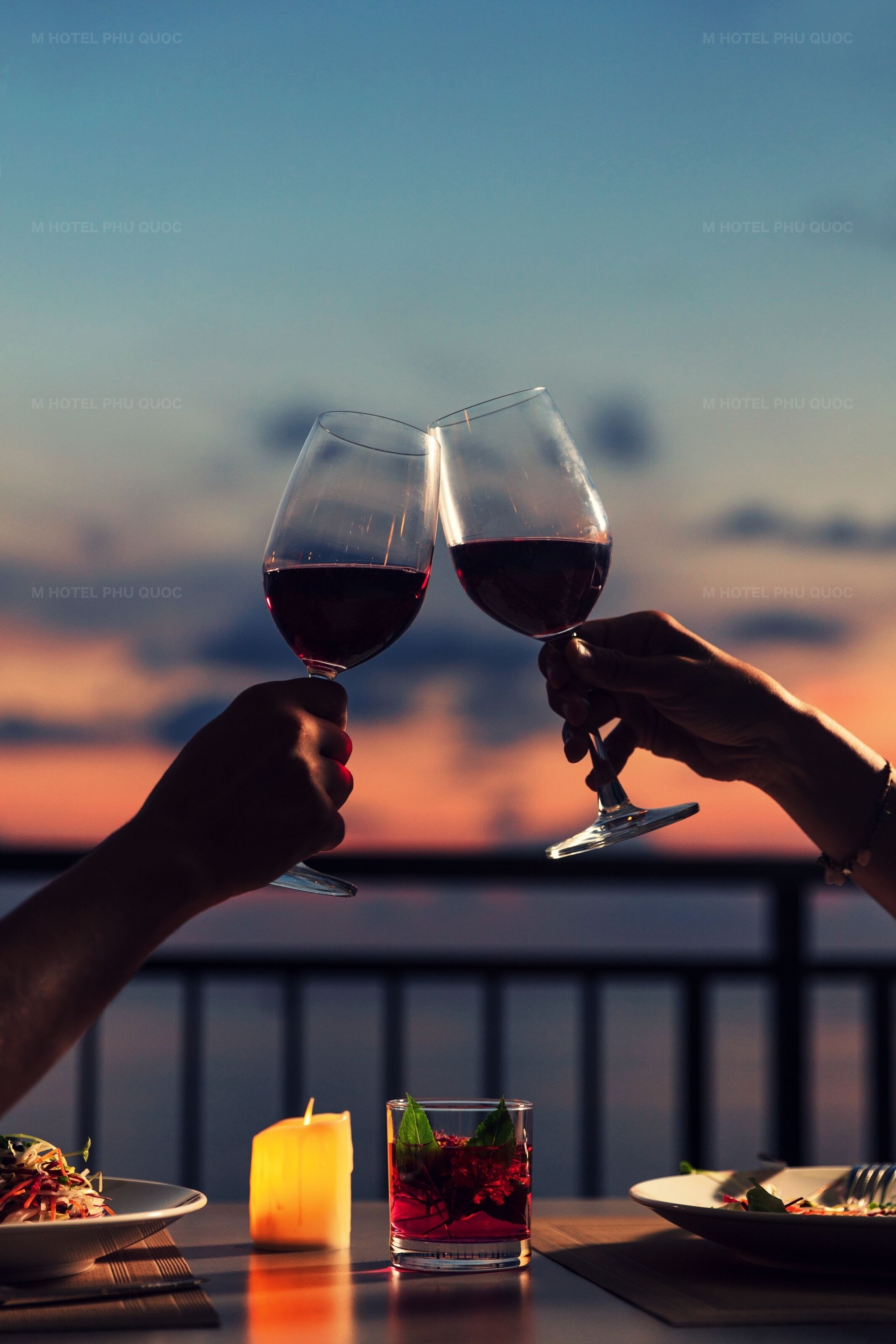 Two hands holding wine glasses toasting with a sunset view in the background.