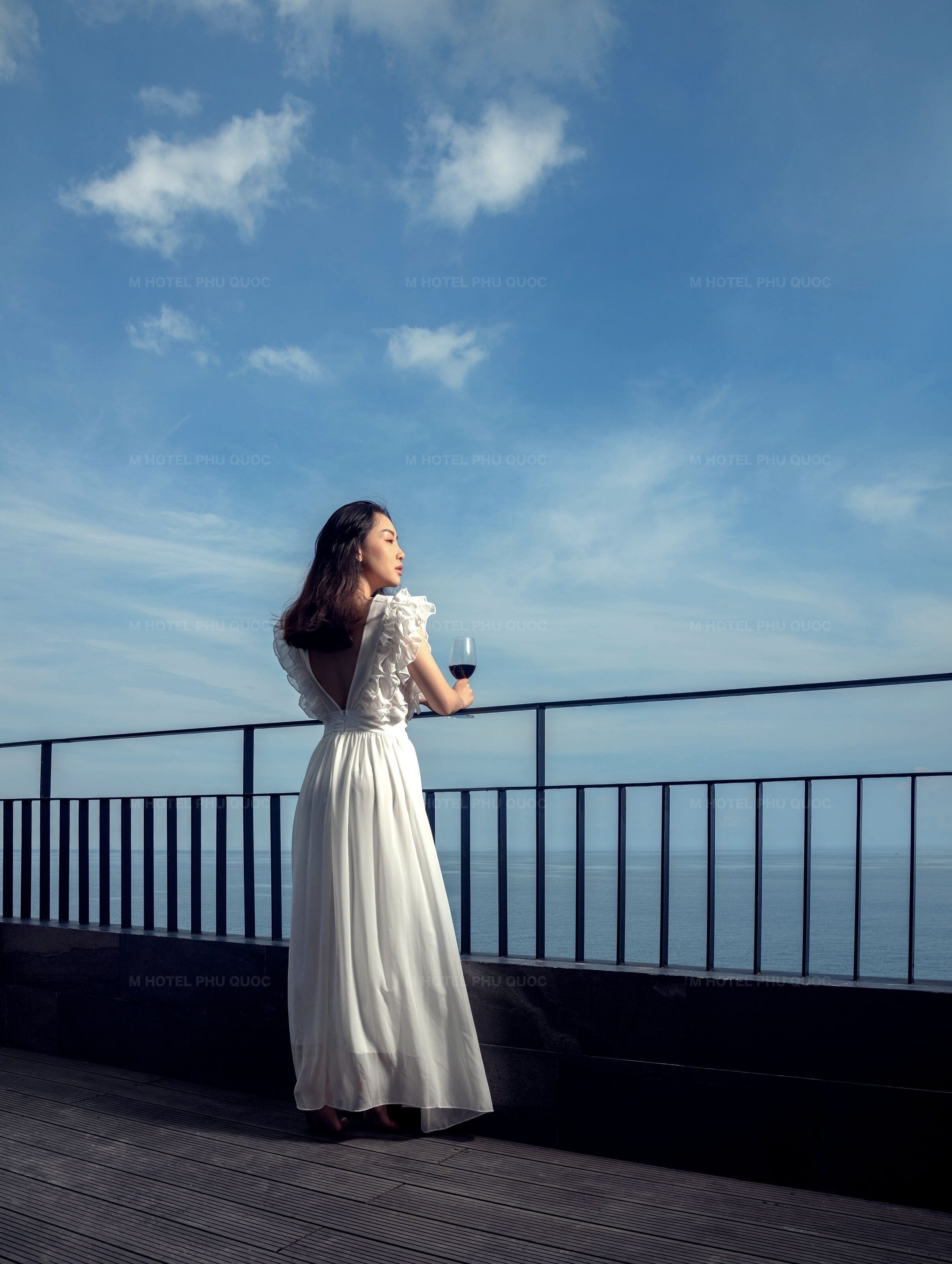 A person in a white dress standing on a balcony with a railing, overlooking a body of water.