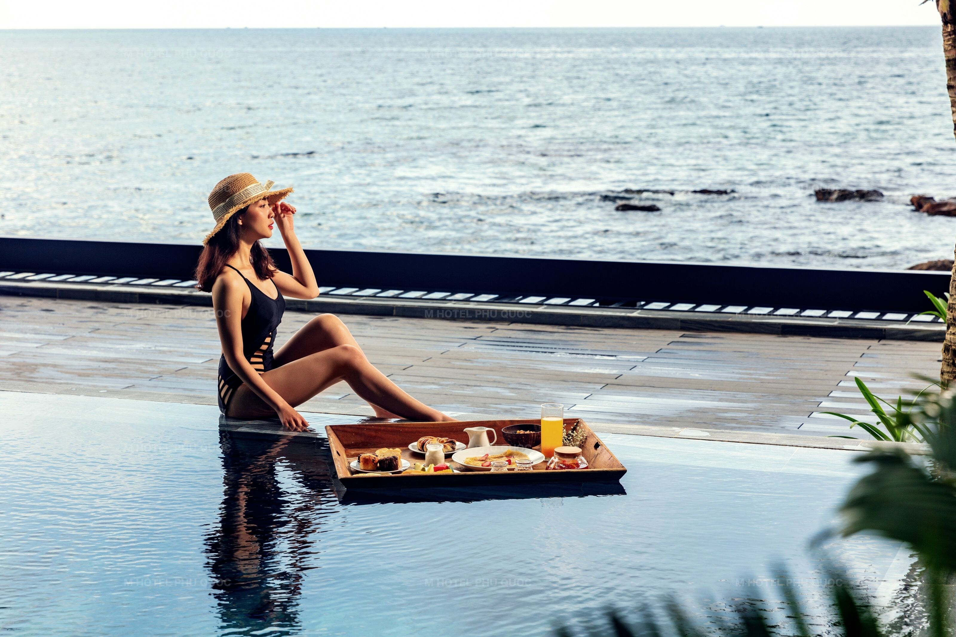 A woman sitting by a pool with a tray of breakfast items.