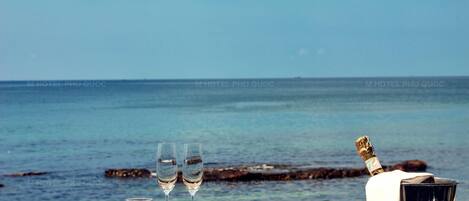 A table with two glasses of champagne, a bottle of champagne, and a bottle of red wine on a deck overlooking the ocean.