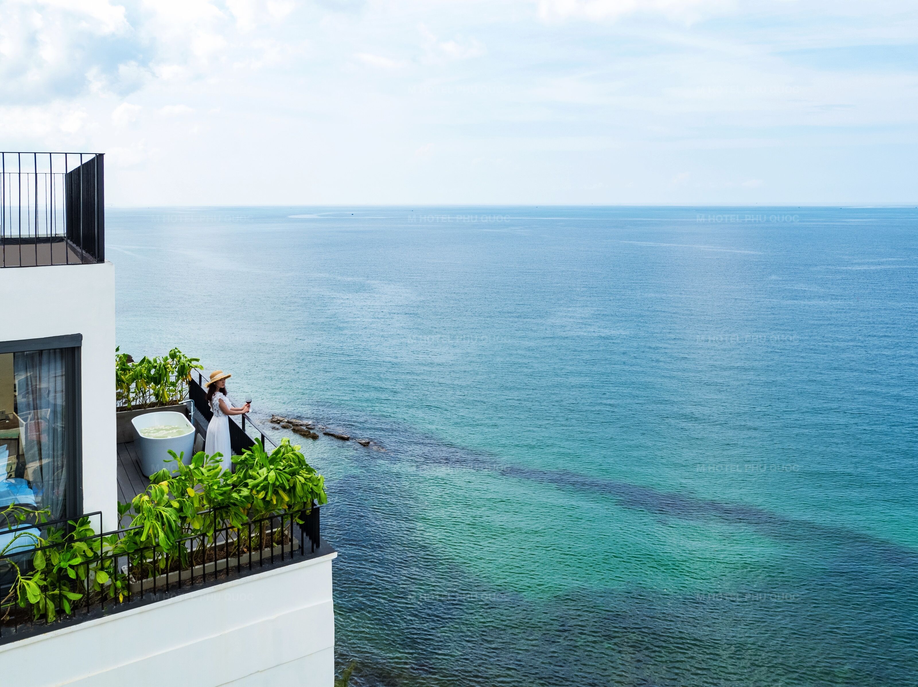 A balcony with a view of the sea, a person in a white dress, and a plant.
